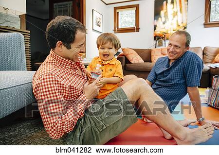Stock Image - Gay fathers playing with baby son in living room. Fotosearch