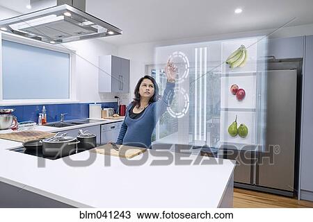 Stock Image - Mixed race woman using holographic screen in kitchen. Fotosearch