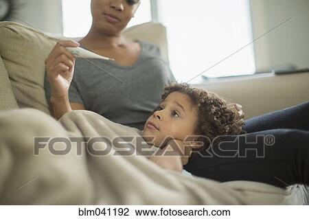 Stock Image - Mother measuring temperature of daughter on sofa. Fotosearch