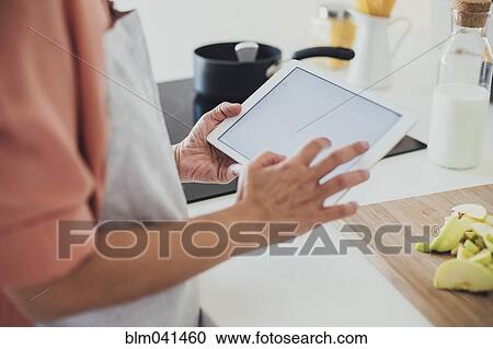 Stock Image - Older Caucasian woman using digital tablet in kitchen. Fotosearch