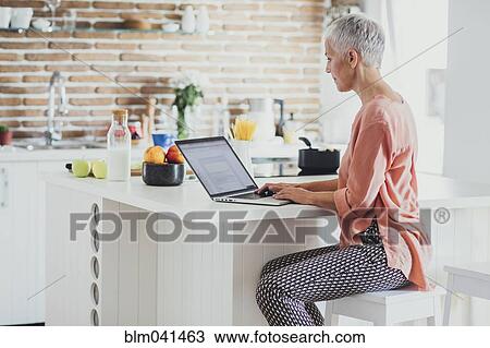 Stock Image - Older Caucasian woman using laptop in kitchen. Fotosearch