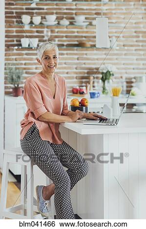 Stock Photograph - Older Caucasian woman using laptop in kitchen. Fotosearch