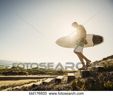 Stock Image - Caucasian man carrying surfboard on beach. Fotosearch