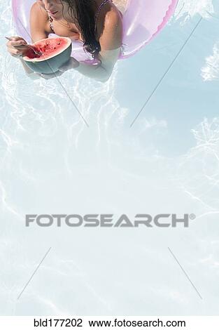 Stock Image - Caucasian woman eating watermelon in swimming pool. Fotosearch