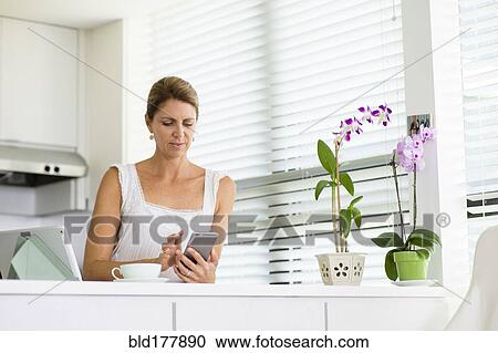 Stock Image - Caucasian woman using cell phone in kitchen. Fotosearch