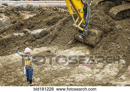 Stock Photo - Caucasian worker directing digger at construction site. Fotosearch