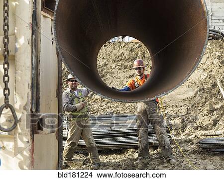 Picture - Caucasian workers hauling pipe at construction site. Fotosearch