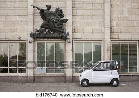 Stock Image - Compact car parked under sculpture on building. Fotosearch
