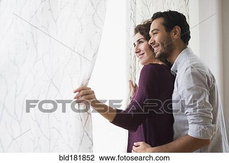 Stock Image - Couple looking out window curtain. Fotosearch