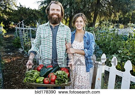Stock Image - Couple standing with basket of vegetables in garden. Fotosearch