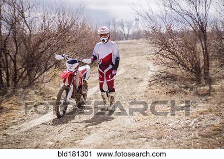 Stock Image - Dirt bike rider standing with motorcycle in rural field. Fotosearch