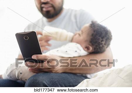 Father bottle feeding baby son and using cell phone View Large Photo Image Stock Photo - Father bottle feeding baby son and using cell phone. Fotosearch