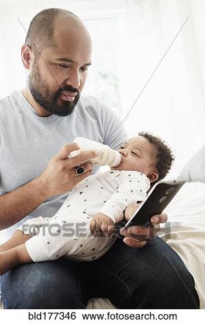 Father bottle feeding baby son and using cell phone View Large Photo Image Stock Photograph - Father bottle feeding baby son and using cell phone. Fotosearch