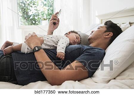 Stock Image - Father using cell phone with sleeping baby son. Fotosearch