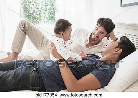 Picture - Gay fathers playing with baby son on bed. Fotosearch