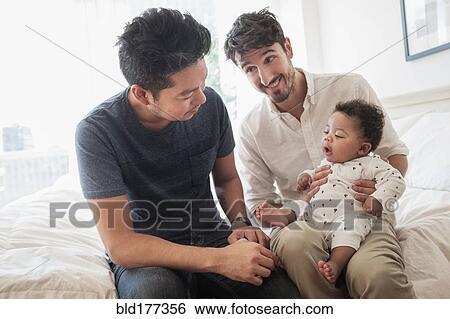Stock Photograph - Gay fathers playing with baby son on bed. Fotosearch