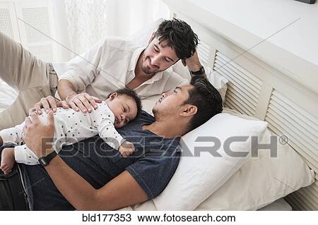Stock Image - Gay fathers relaxing with baby son on bed. Fotosearch
