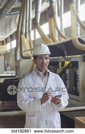 Stock Image - Hispanic technician using cell phone factory. Fotosearch