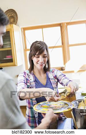 Stock Image - Hispanic woman serving boyfriend in kitchen. Fotosearch