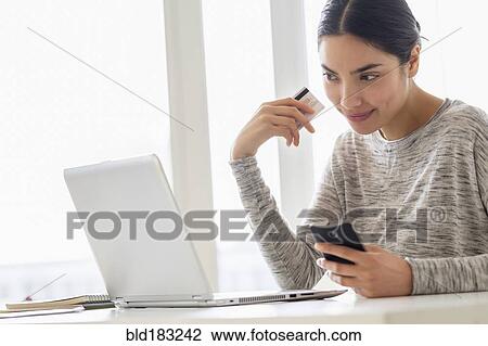 Stock Image - Hispanic woman shopping on cell phone and laptop. Fotosearch