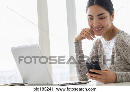 Stock Image - Hispanic woman using cell phone and laptop. Fotosearch