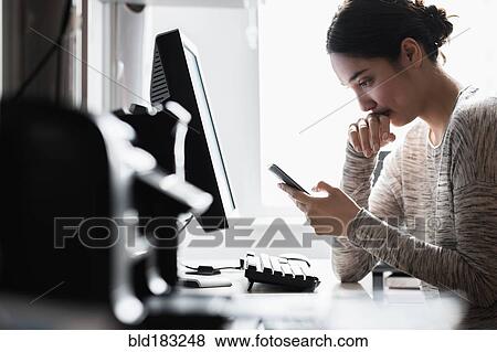 Stock Photo - Hispanic woman using cell phone at computer. Fotosearch