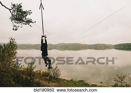 Stock Image - Man hanging on rope swing at remote lake. Fotosearch