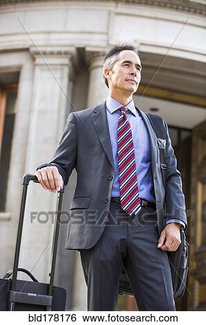 Stock Photograph - Mixed race businessman rolling luggage outdoors. Fotosearch