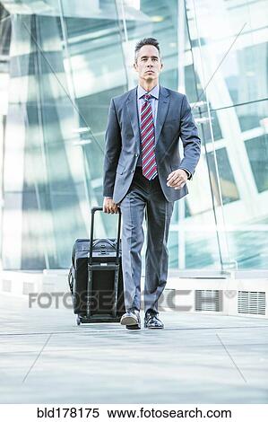 Stock Photography - Mixed race businessman rolling luggage outdoors. Fotosearch