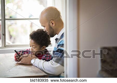 Stock Photography - Mixed race father and son using cell phone at table. Fotosearch