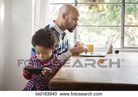 Stock Image - Mixed race father and son using technology at table. Fotosearch