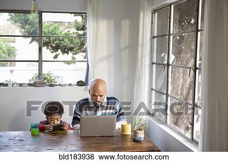 Stock Photo - Mixed race father and son using technology at table. Fotosearch