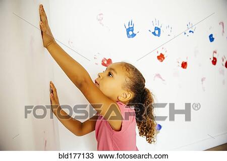 Stock Image - Mixed race girl making hand prints on wall. Fotosearch