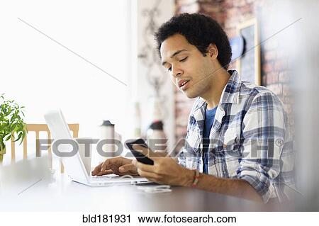 Stock Image - Mixed race man multitasking in coffee shop. Fotosearch