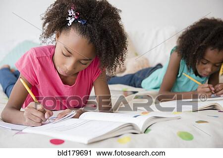 Stock Image - Mixed race sisters doing homework on bed. Fotosearch