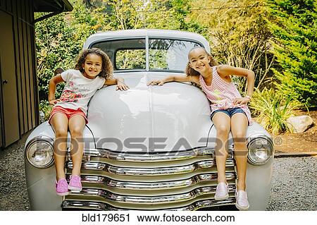 Stock Image - Mixed race sisters sitting on vintage car. Fotosearch