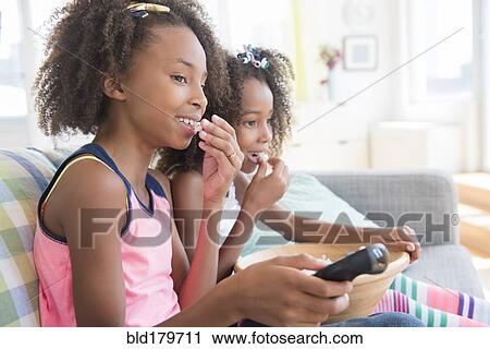 Stock Image - Mixed race sisters watching television on sofa. Fotosearch