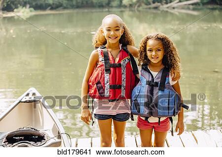Stock Image - Mixed race sisters wearing life jackets at lake. Fotosearch
