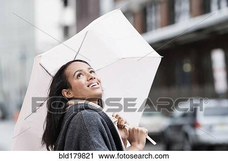Stock Image - Mixed race woman carrying umbrella outdoors. Fotosearch