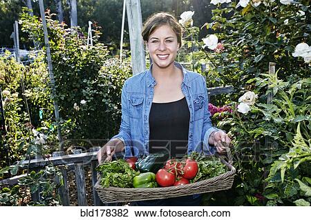 Stock Image - Mixed race woman holding basket of vegetables in garden. Fotosearch