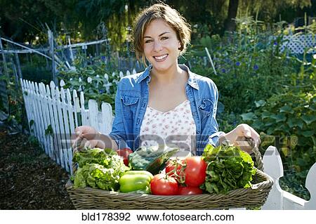 Stock Image - Mixed race woman holding basket of vegetables in garden. Fotosearch