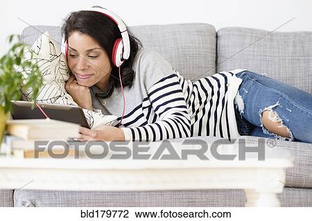 Stock Image - Mixed race woman listening to headphones on sofa. Fotosearch