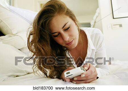 Stock Image - Mixed race woman using cell phone on bed. Fotosearch