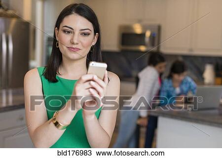 Stock Image - Mother using cell phone in kitchen. Fotosearch