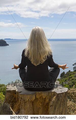 Stock Image - Older Caucasian woman meditating on cliff near ocean. Fotosearch