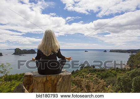 Stock Image - Older Caucasian woman meditating on cliff near ocean. Fotosearch
