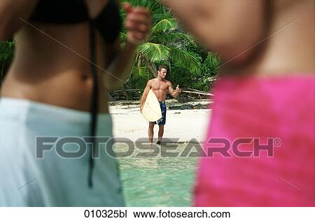 Stock Image - blurred image of two women in foreground watching man with surfboard. Fotosearch