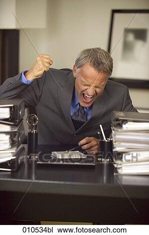 Stock Image - businessman behind his desk raising his fist in anger. Fotosearch