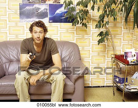 Stock Image - caucasian male sitting on grey sofa zapping. Fotosearch