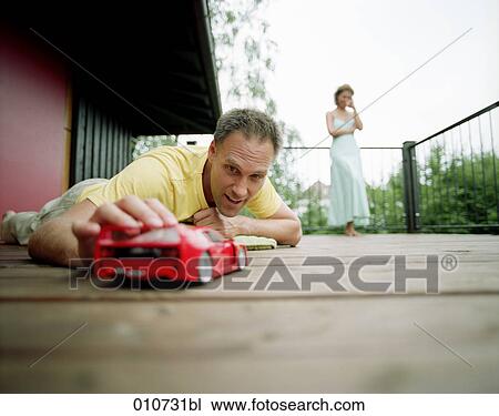 Stock Image - man playing with red toy car while woman is standing in the background. Fotosearch